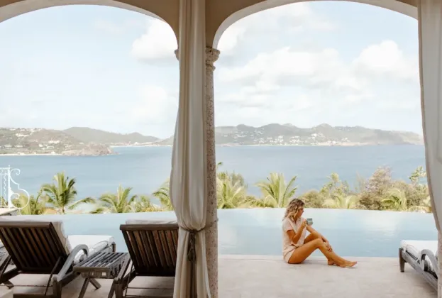 lady sitting next to infinity edge pool holding a coffee mug and views of palm trees and ocean in background
